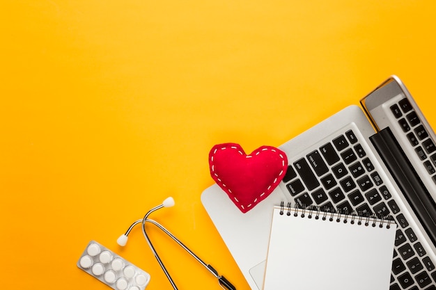 Free Photo high angle view of laptop; spiral notepad; blister packed medicine; stethoscope; stitched heart shape above yellow backdrop