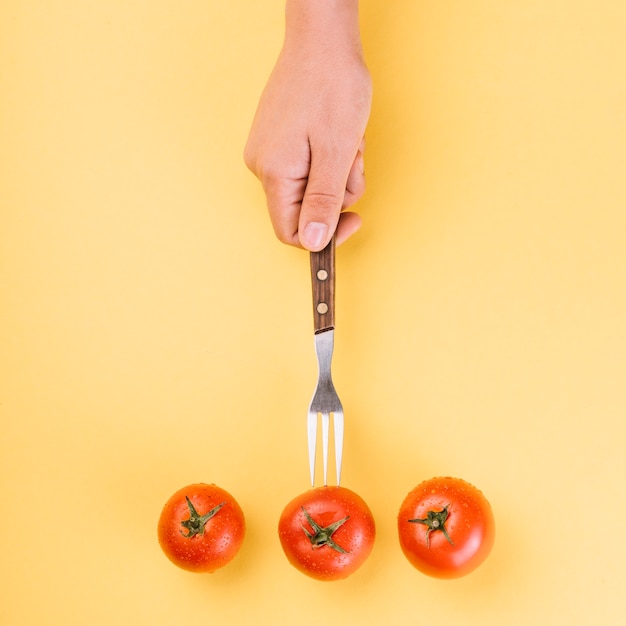 Free Photo high angle view of a person's hand inserting fork in red tomato on yellow background