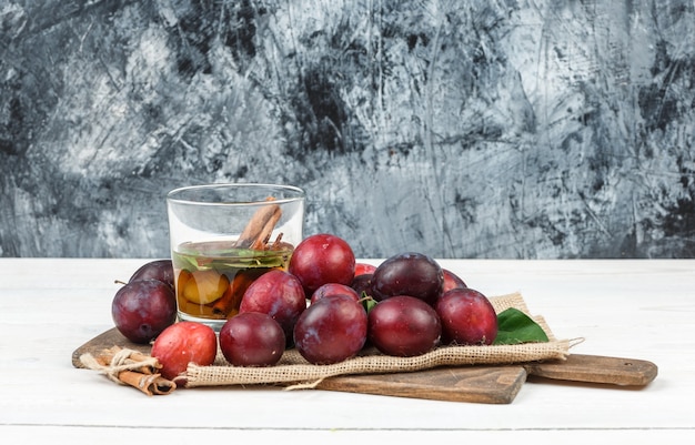 High angle view plums and detox water on cutting board with leaves and a piece of sack on wooden board and dark blue marble surface. horizontal