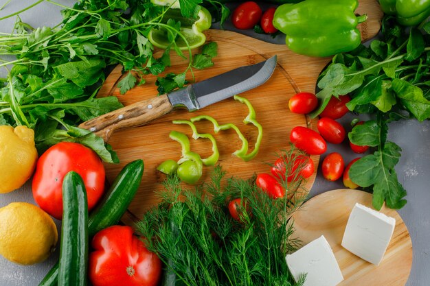 High angle view sliced green pepper on cutting board with tomatoes, salt, cheese, lemon, greens, knife on gray surface