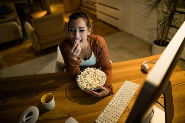 Free photo high angle view of smiling woman watching something on the internet while eating popcorn at night at home