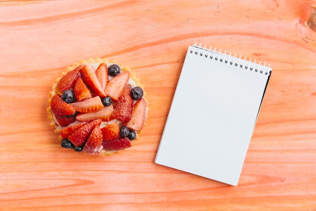 High angle view of strawberry tart and spiral notepad on wooden surface