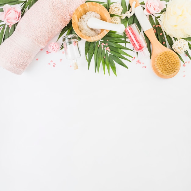 Free Photo high angle view of towel; salt; loofah; leaves; bottle; brush and fake flowers on white surface