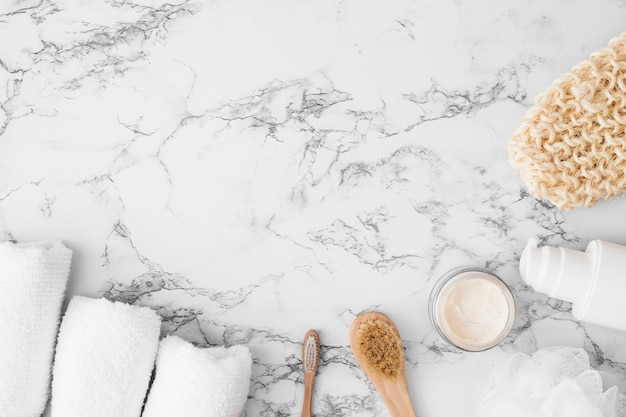 High angle view of towels; brush; moisturizing cream; sponge; cosmetic bottle and scrub glove on marble surface