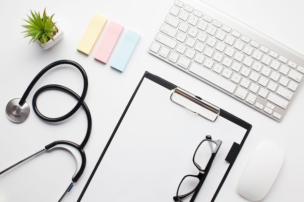 High angle view of wireless devices; spectacles on clipboard; stethoscope and adhesive notes over white table