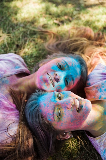 Free Photo high angle view of young women with holi color on their face lying on lawn looking at camera