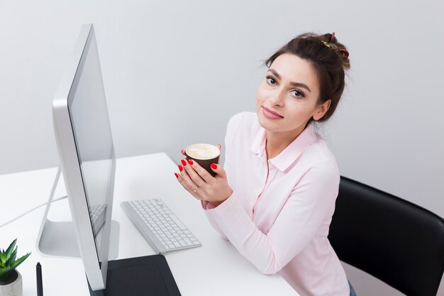 High angle of woman posing with cup of coffee at desk next to computer