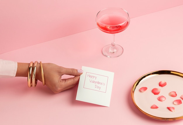 High angle of woman's hand holding valentines day card next to glass and rose petals