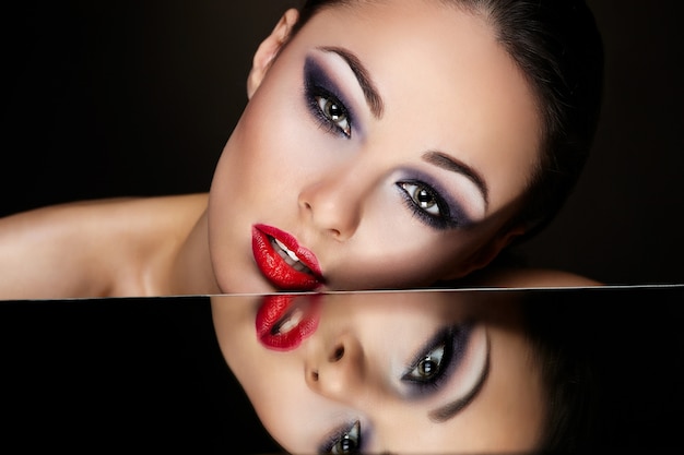 Free photo high fashion look.glamour fashion portrait of beautiful sexy brunette girl with bright makeup and red lips and her reflection in mirror table on dark