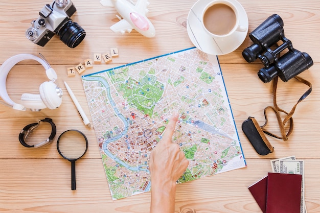 Hiker pointing at location on map with cup of tea and traveler accessories on wooden background