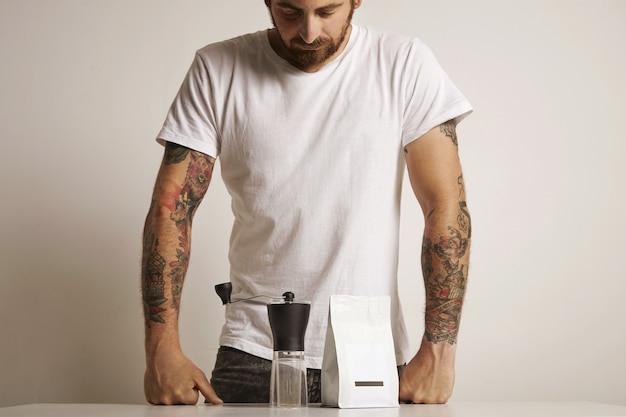 Free Photo hip tattooed barista in plain white t-shirt looking down at a small manual burr grinder and unlabeled white bag with coffee beans