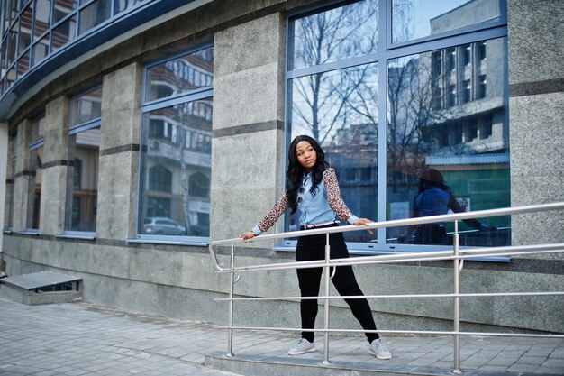 Hipster african american girl wearing jeans shirt with leopard sleeves posing at street against modern office building with blue windows