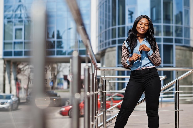 Hipster african american girl wearing jeans shirt with leopard sleeves posing at street against modern office building with blue windows