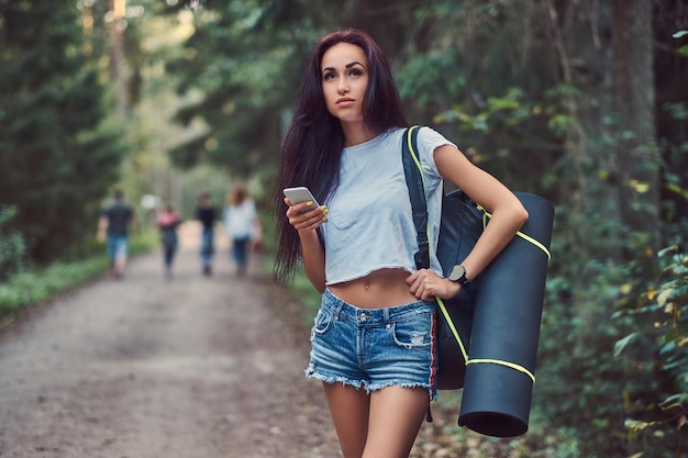 Free photo hipster girl dressed in a shirt and shorts with tourist mat and backpack holds a smartphone and looking away while standing in a summer forest.