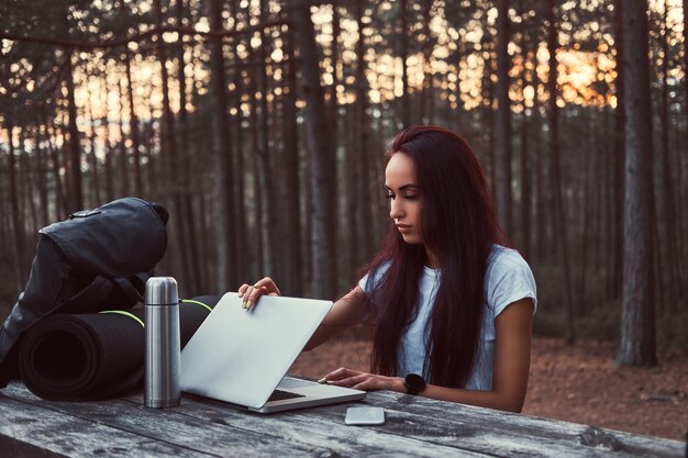 Free photo hipster girl opening a laptop for working on a wooden bench while having a break in a beautiful autumn forest.