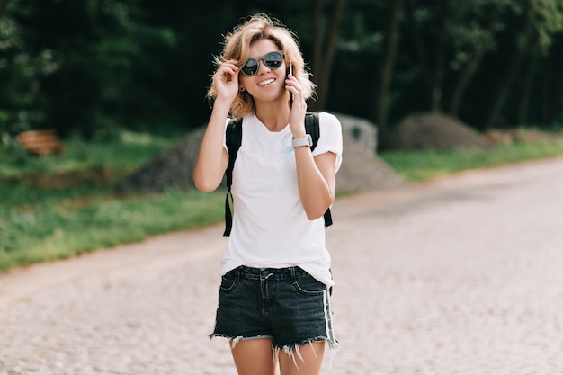 Hipster young girl with backpack enjoying trip. Tourist traveler over valley landscape view. Female traveler walking on the road and talking on the phone