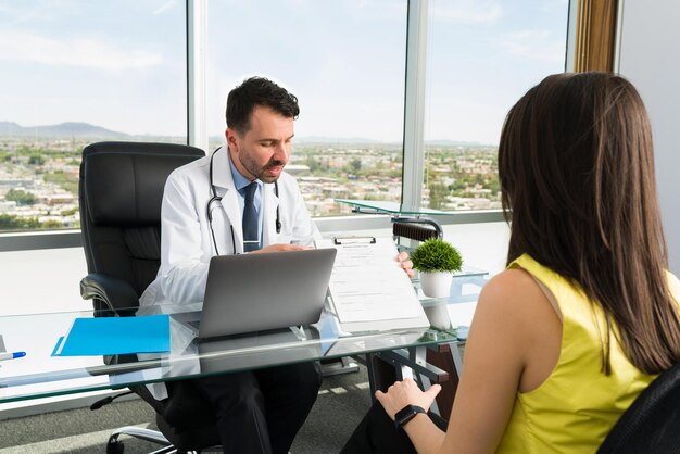 Hispanic doctor talking with a female patient at his office and asking her to fill a medical form with her symptoms to find her a treatment