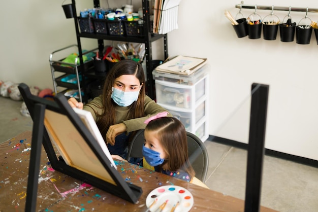 Hispanic female teacher is instructing a little girl easy painting techniques in a blank canvas during an art class. Young woman and elementary kid wearing face masks