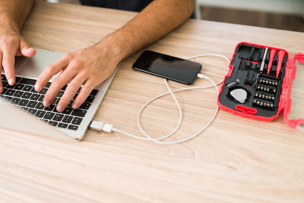 Free photo hispanic male technician typing on laptop and connecting a smartphone to the computer to make a data backup at his repair shop