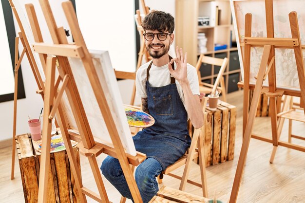 Hispanic man with beard at art studio doing ok sign with fingers, smiling friendly gesturing excellent symbol