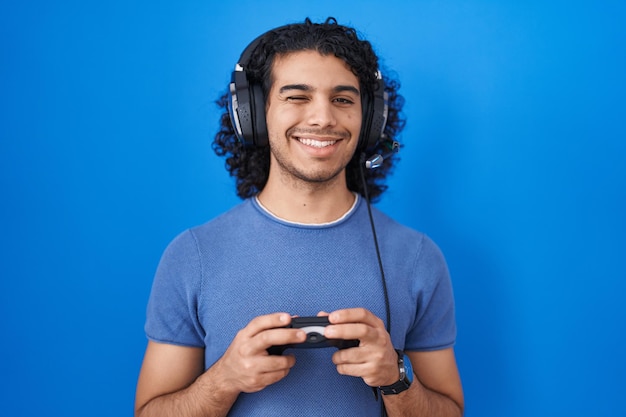 Hispanic man with curly hair playing video game holding controller winking looking at the camera with sexy expression, cheerful and happy face.