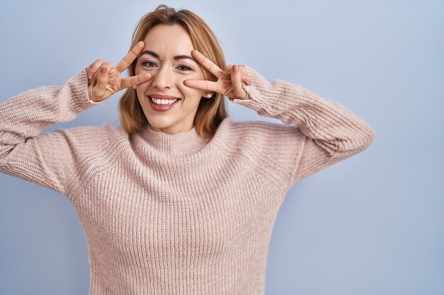 Hispanic woman standing over blue background doing peace symbol with fingers over face smiling cheerful showing victory