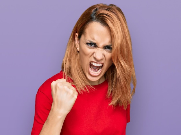 Free photo hispanic young woman wearing casual red t shirt angry and mad raising fist frustrated and furious while shouting with anger. rage and aggressive concept.