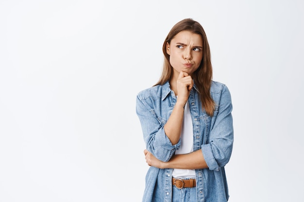 Hmm something strange. Thoughtful young woman having doubts, frowning and pouting while thinking, looking at upper left corner pensive, standing on white