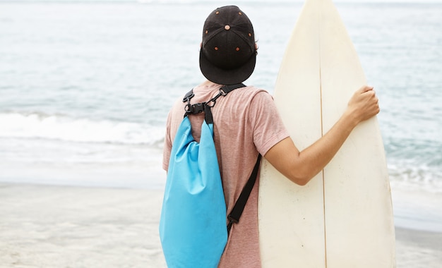 Free photo hobby, leisure and summer vacations. back shot of stylish young surfer wearing snapback and backpack going to ride waves while having holidays in tropical country