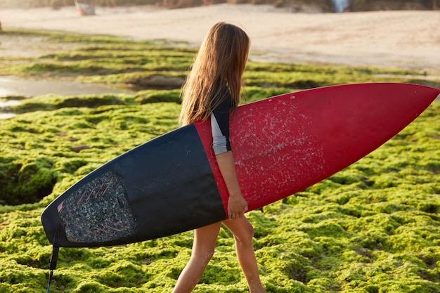 Free photo hobby and sport concept. active female surfer carries surf board, walks on coast during summer holidays, wants to hit ocean waves, has recreation in paradise place, poses alone. horizontal shot