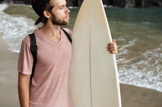 Free Photo hobby, watersports and active lifestyle concept. young surfer wearing baseball cap backwards standing alone on sandy beach and looking at sea with thoughtful expression