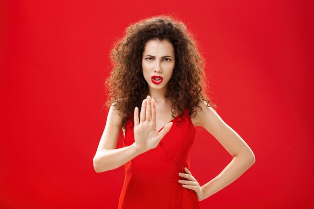 Hold on stop. Intense serious-looking displeased adult woman with curly hair in stylish red dress frowning raising palm in no gesture refusing giving negative answer, prohibiting come closer.