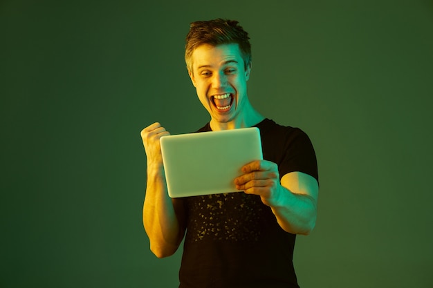 Holding tablet, celebrating win in bet or game. Caucasian man's portrait on green studio background in neon light. Beautiful male model. Concept of human emotions, facial expression, sales, ad.