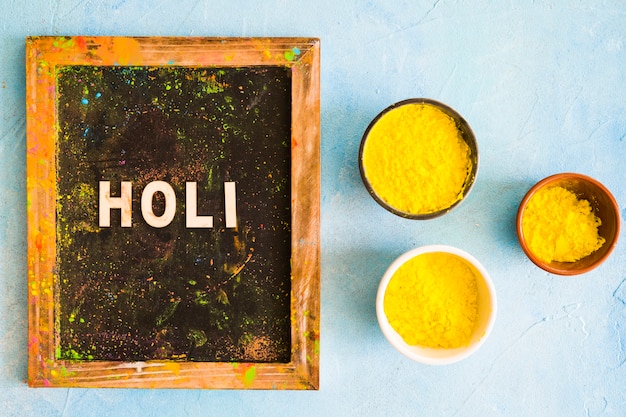 Holi text on messy wooden slate with yellow holi colored bowls on painted backdrop