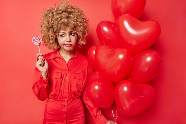 Free photo holiday concept sad dissatisfied young woman with curly hair wears dress holds lollipop and bunch of inflated heart balloons isolated over vivid red background feels very tired after party