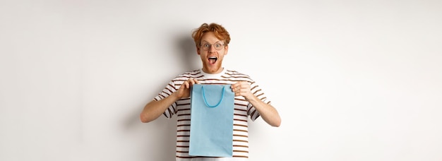 Holidays and celebration concept surprised redhead man receiving present inside shopping bag looking