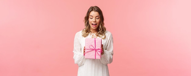 Holidays celebration and women concept Portrait of surprised charming young blond girl receive surprise gift holding present in pink box look at it amused curious whats inside studio background