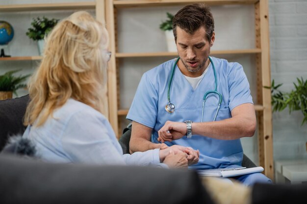 Home caregiver checking pulse of his patient and looking at wristwatch while being in a home visit