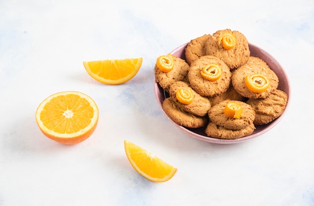 Homemade cookies in pink bowl and orange slices over white table.