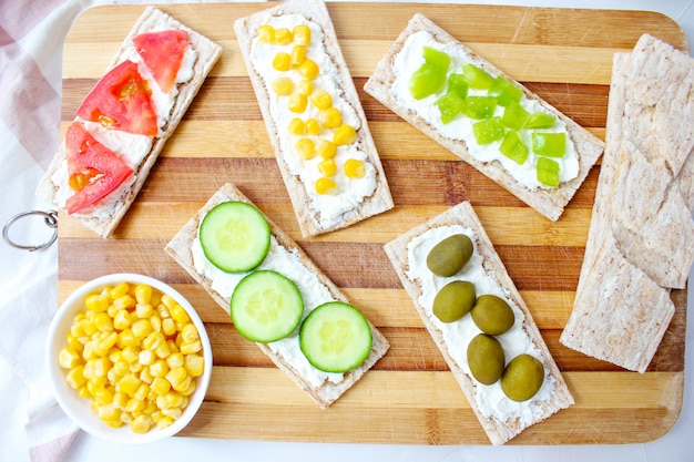 Free photo homemade crispbread toast with cottage cheese and green olives, slices of cabbage, tomatoes, corn, green pepper on cutting board. healthy food concept, top view. flat lay