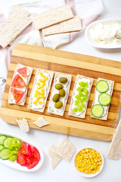 Free photo homemade crispbread toast with cottage cheese and green olives, slices of cabbage, tomatoes, corn, green pepper on cutting board. healthy food concept, top view. flat lay