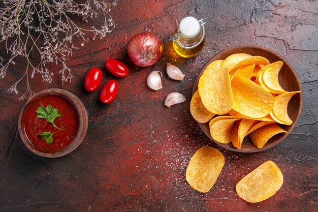 Homemade delicious crispy potato chips inside and outside of brown pot oil bottle ketchup tomatoes garlic onion on dark background