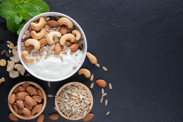 Homemade granola or muesli, bowl of oat granola with yogurt, almond, Cashew nuts, mint and nuts on the black rock board for healthy breakfast, copy space. Healthy breakfast menu concept.