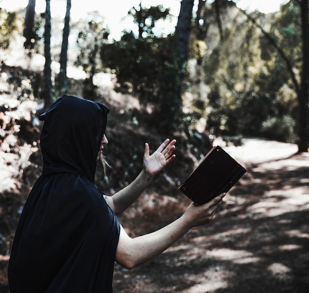 Free Photo hooded woman with book conjuring in sunlit woodland