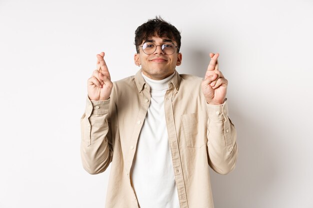 Hope. Happy young man making wish and smiling, looking hopeful at camera as waiting for dream come true, cross fingers for good luck, standing on white background