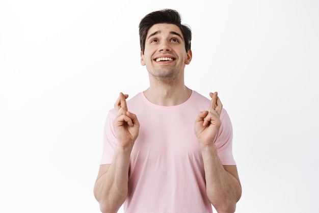 Free photo hope and luck smiling young man cross fingers and look up praying asking god for make wish come true anticipating positive news standing over white background