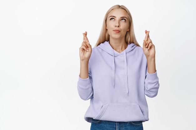 Free Photo hopeful blond girl cross fingers for good luck and looking at upper left corner, praying, making wish for dream come true, standing on white