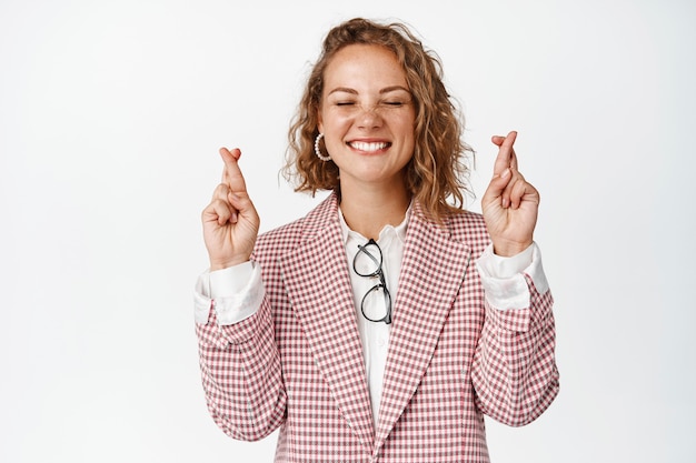 Hopeful business woman close eyes and smiles, cross fingers for good luck, anticipating good news, standing on white