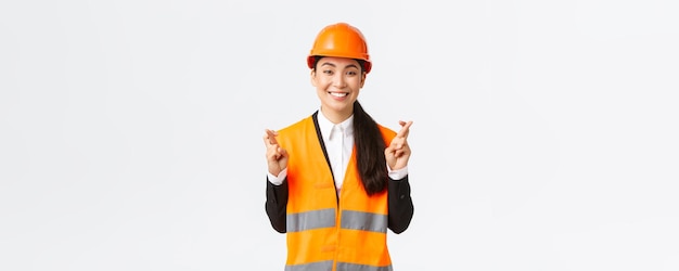 Hopeful and optimistic smiling asian female engineer construction manager in safety helmet having faith in project cross fingers good luck and looking positive standing white background