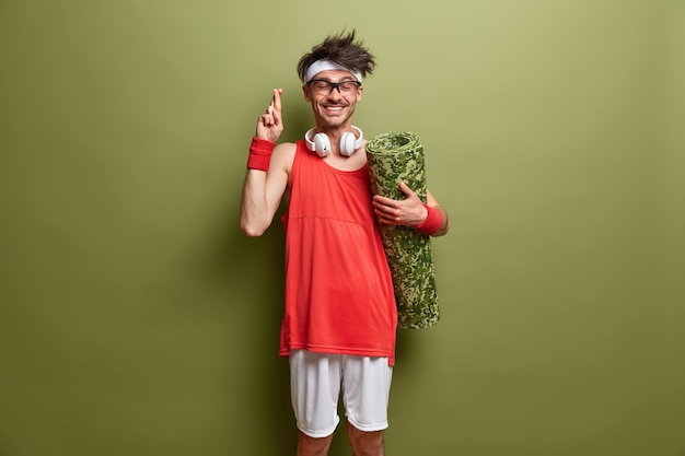 Free Photo hopeful positive man crosses fingers and believes in successful training, prepares for workout, holds karemat, wears sport clothes, has cheerful expression isolated on green wall. time for sport